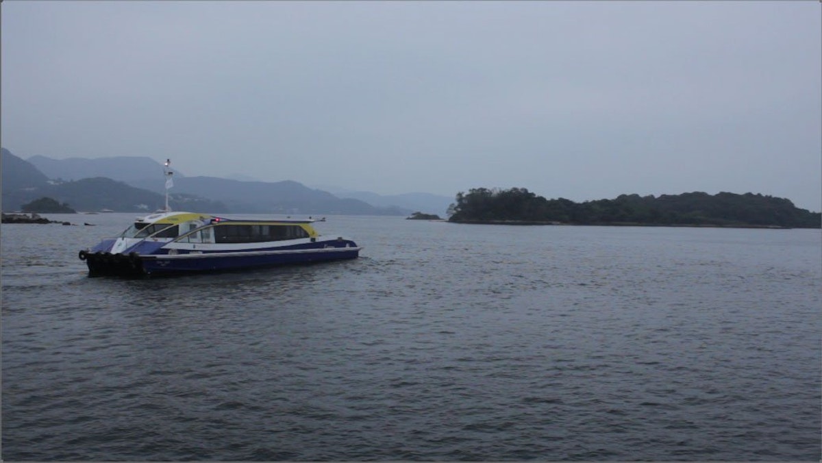 A short and long ferry floats on a body of water on a dark and cloudy day. The ferry sits on water next to a small island in the background.
